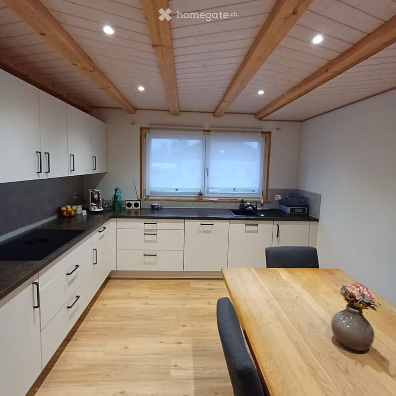 Modern kitchen with white cabinets, black countertops, and a wooden dining table with chairs. The space features a window above the sink and a sleek, contemporary design.