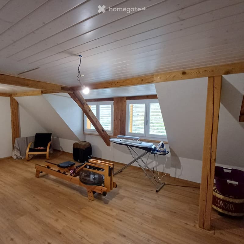 An attic room with wooden floors, slanted ceilings, and exposed wooden beams. Features include a chair, ironing board, iron, exercise machine, and a small window with blinds.
