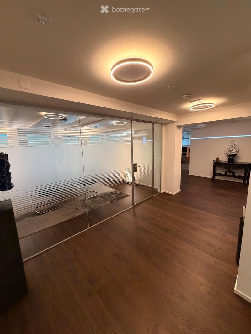 Modern hallway with wooden flooring, glass partitions, and recessed lighting. Features a small seating area with a table and vase in the background.