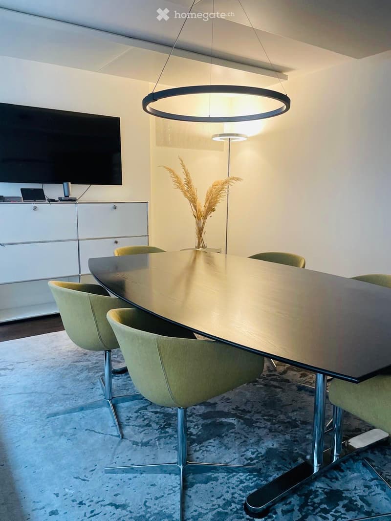 Modern conference room with a large glass table, green chairs, a hanging circular light fixture, a flat-screen TV, and a cabinet with electronic devices.