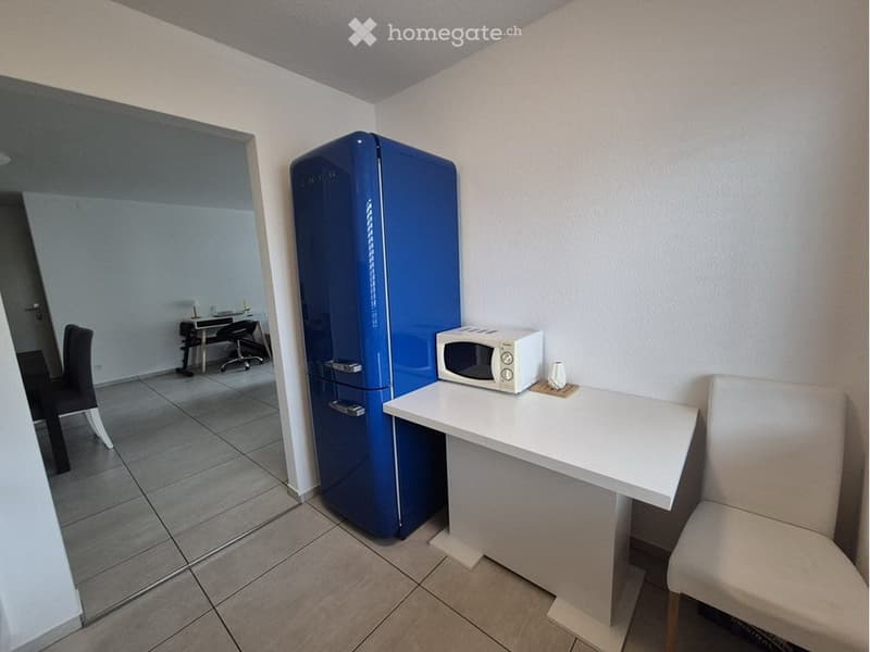 A small, modern kitchen area with a blue refrigerator, a white microwave on a counter, a small table with a chair, and a reflection of a dining area in the mirror.