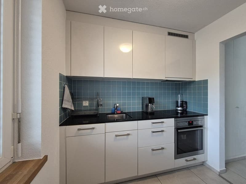 Modern kitchen with white cabinets, black countertops, and stainless steel appliances including a sink, oven, coffee maker, and blender. Blue tiled backsplash and a small window above the sink.