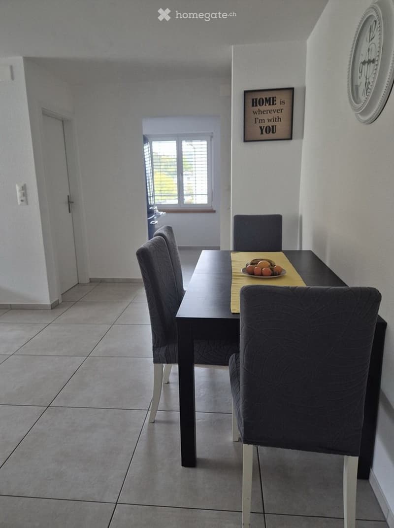 Dining room with a dark wooden table, four grey upholstered chairs, a fruit bowl, and a clock on the wall.