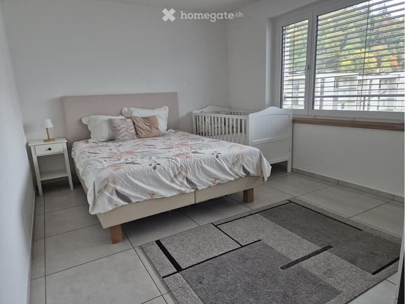 Bedroom with a large bed, nightstand, lamp, baby crib, and a grey rug on the floor.