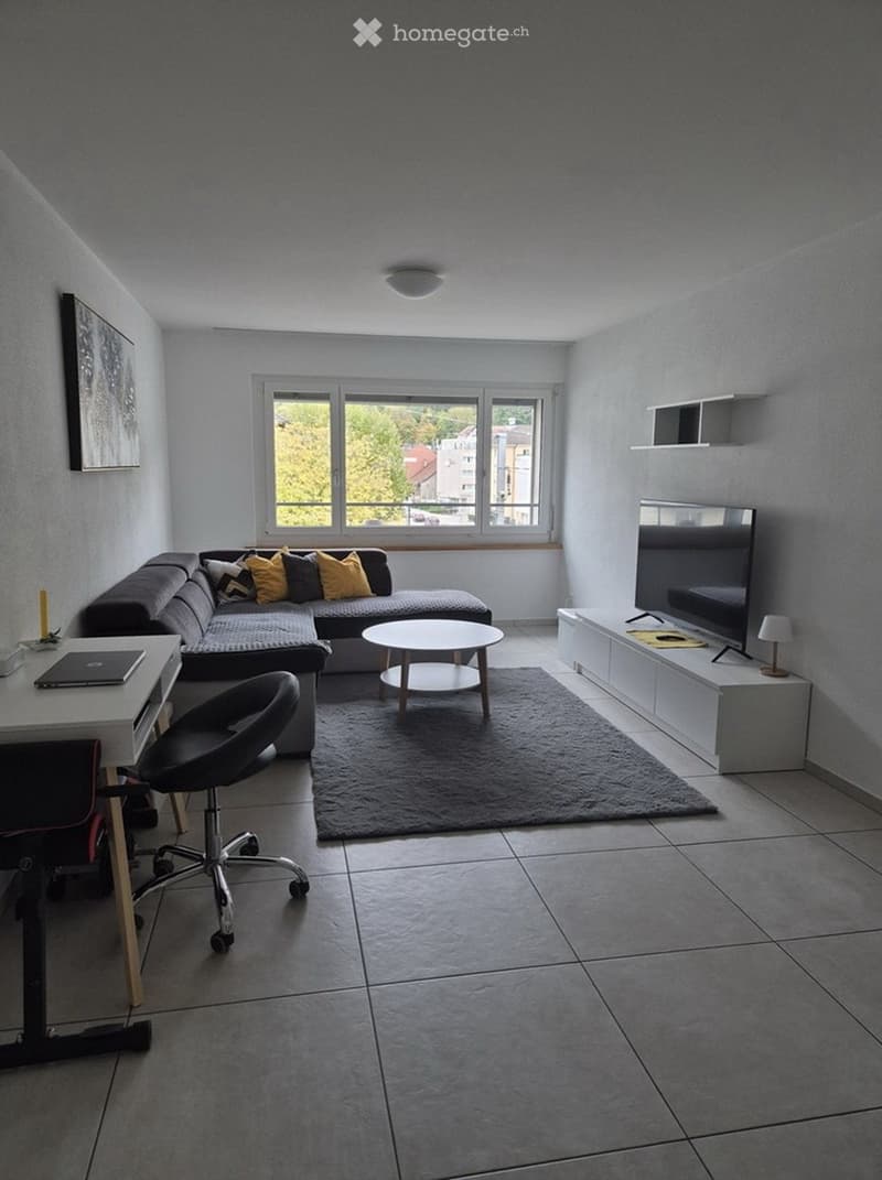 Modern living room with a grey sectional sofa, coffee table, round side table, TV stand, and a desk with a laptop. The room features large windows, a grey rug, and a tiled floor.
