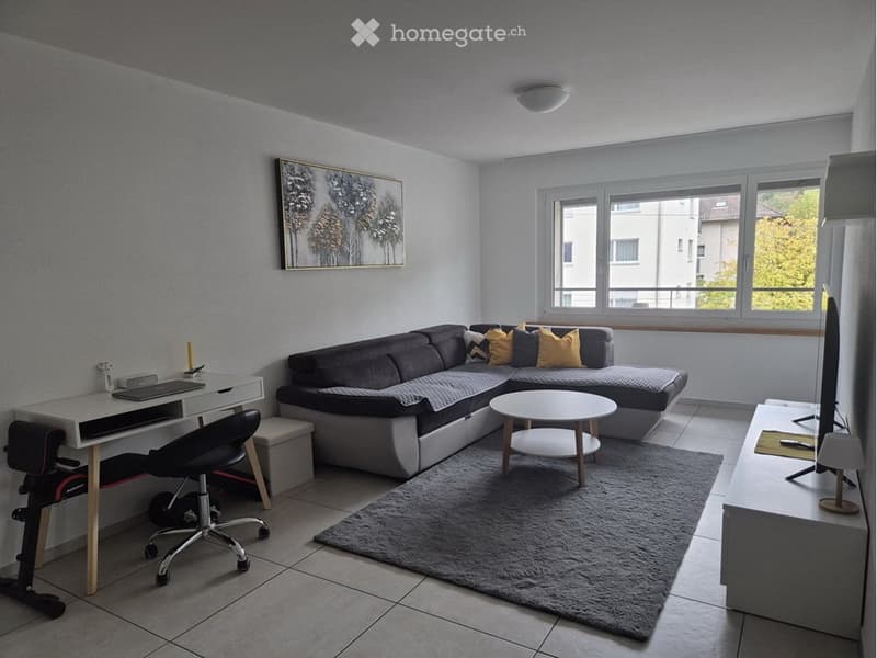 Modern living room with a grey sectional sofa, coffee table, desk with laptop, chair, and a TV screen. Large windows provide natural light.