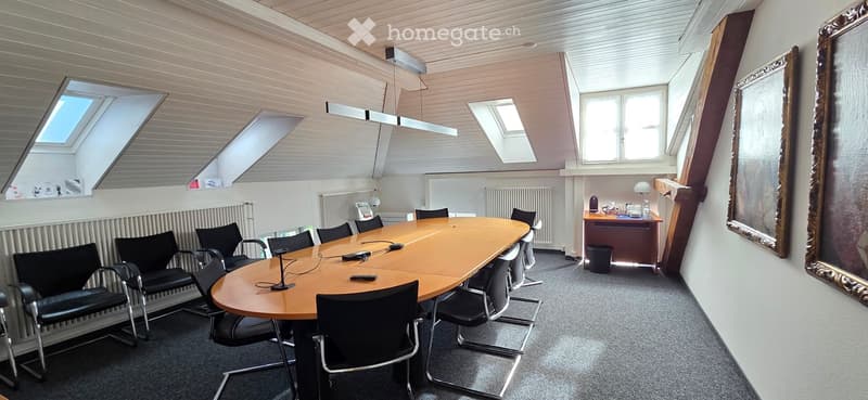 Conference room with a large wooden table surrounded by black chairs, a small desk with a lamp, and a large mirror on the wall. The room has skylights and a modern design.