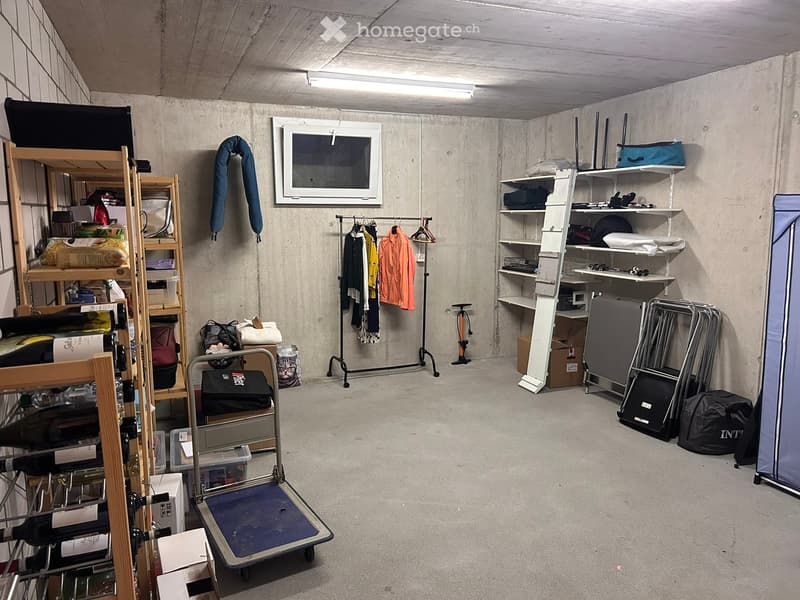 Storage room with concrete walls and ceiling, containing shelves, clothing rack, luggage, camping gear, and a wine rack.
