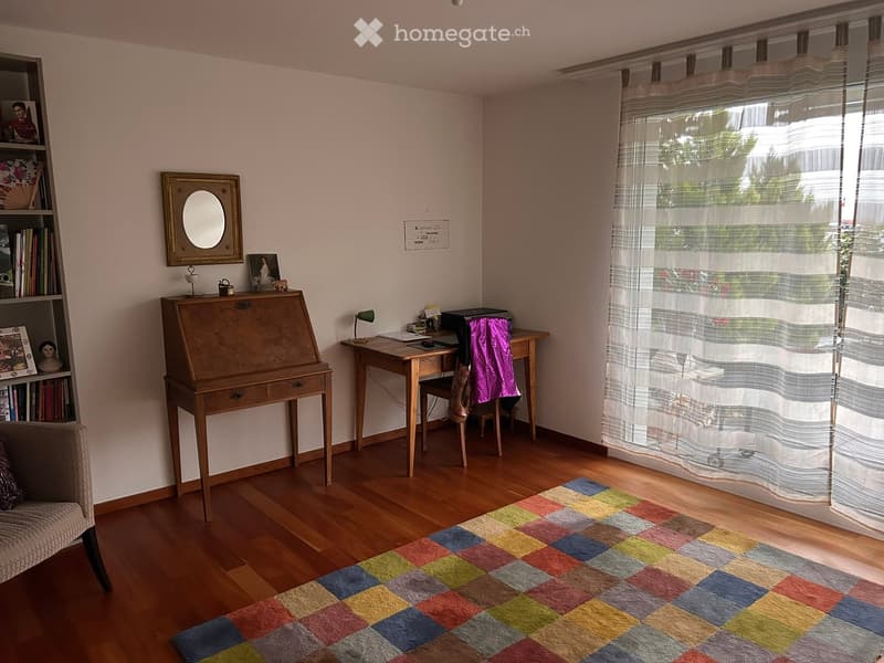 A cozy, well-furnished room with wooden flooring, a colorful checkered rug, a wooden desk, a bookshelf, a mirror, and a sofa.