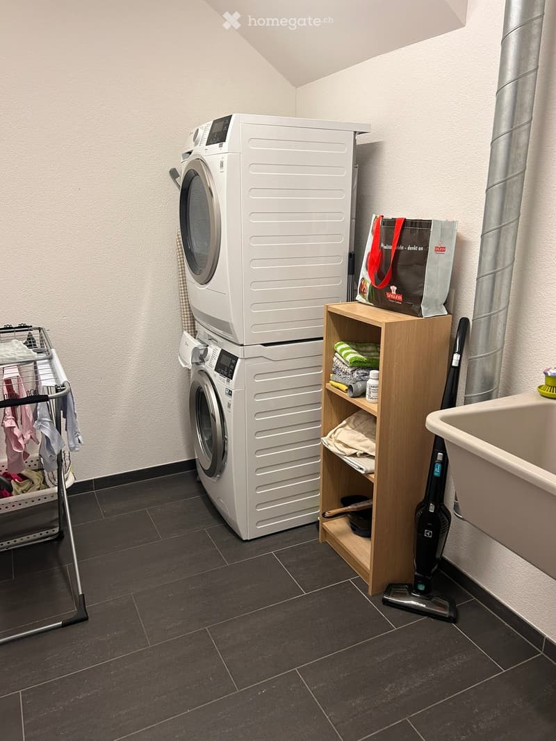 Bathroom with a washing machine and dryer stacked, a wooden shelf with towels and cleaning supplies, a vacuum cleaner, a laundry basket, and a bathtub.