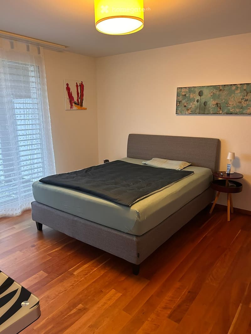 Modern bedroom with wooden flooring, a grey upholstered bed, a nightstand with a lamp, and two picture frames on the wall.