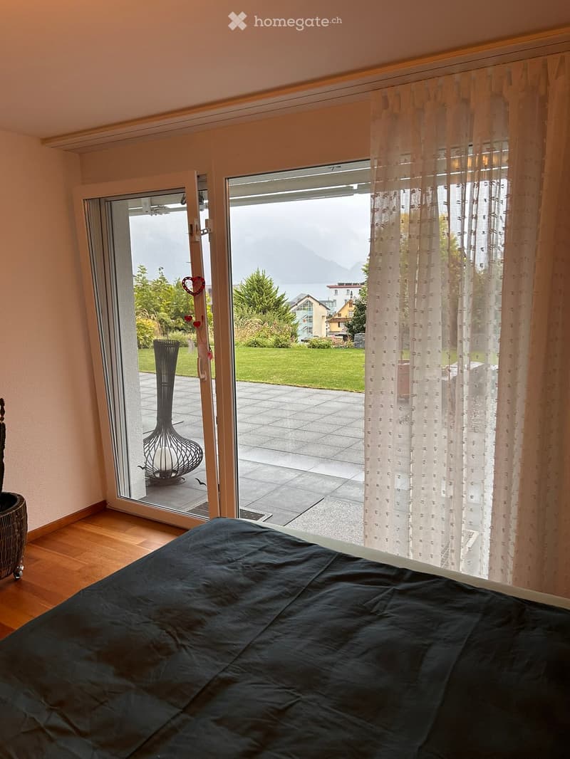 Bedroom with wooden flooring, large window with sheer curtains, and a view of a garden and mountains.