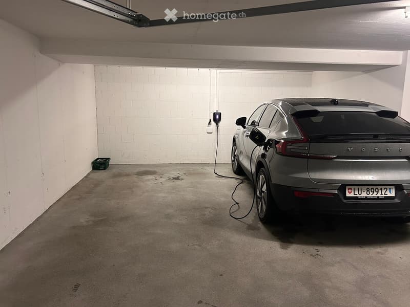 Underground parking space with a single car parked and connected to a charging station. The area has concrete flooring and white brick walls.