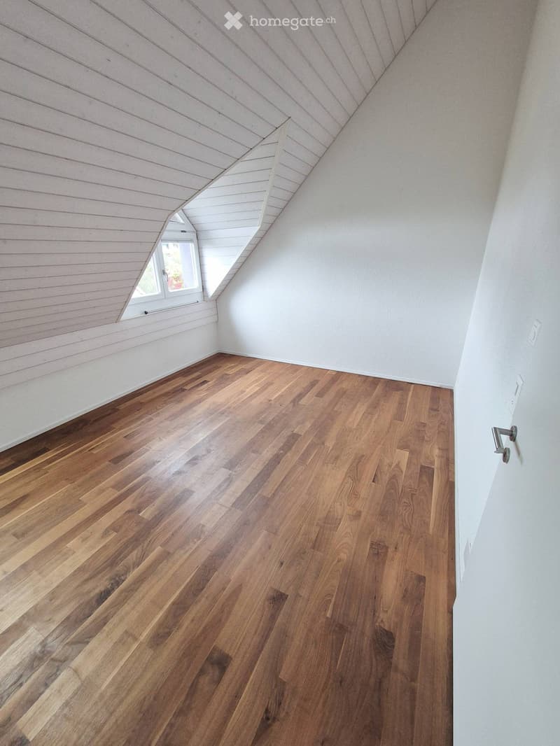Empty attic room with wooden flooring, white walls, and a slanted wooden ceiling. A small window with white frame is visible on the left side.