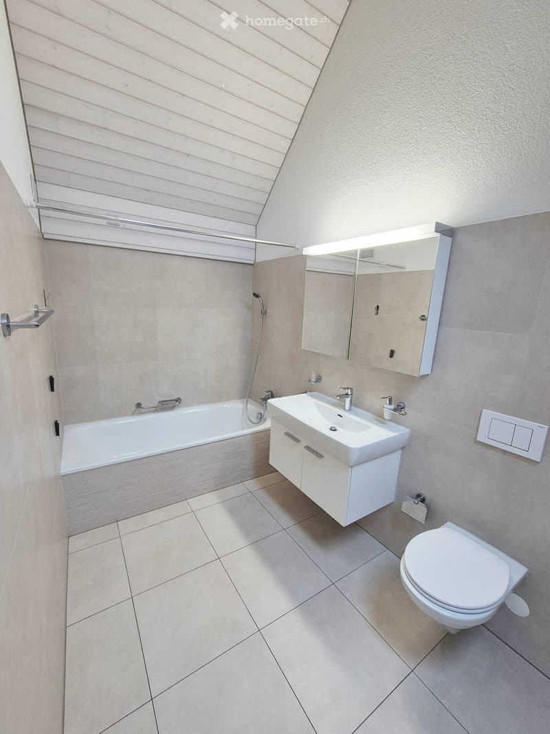 Modern bathroom with a bathtub, sink, toilet, and mirror. Light-colored tiles and a sleek design.