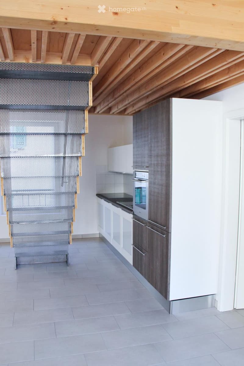 Modern kitchen with white and dark grey cabinets, a built-in oven, and a sleek countertop. The space features a wooden ceiling and a metal staircase leading upwards.