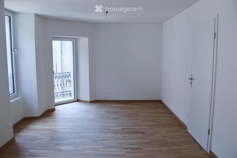 Empty room with wooden flooring, white walls, and a door leading to a balcony with metal railings.