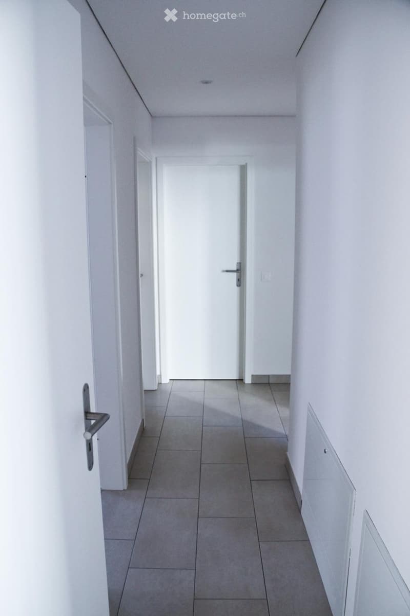 A clean, modern hallway with white walls, tiled floor, and a closed door at the end.