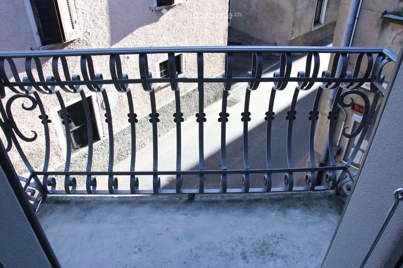Balcony with ornate black metal railing, overlooking a narrow street with adjacent buildings.