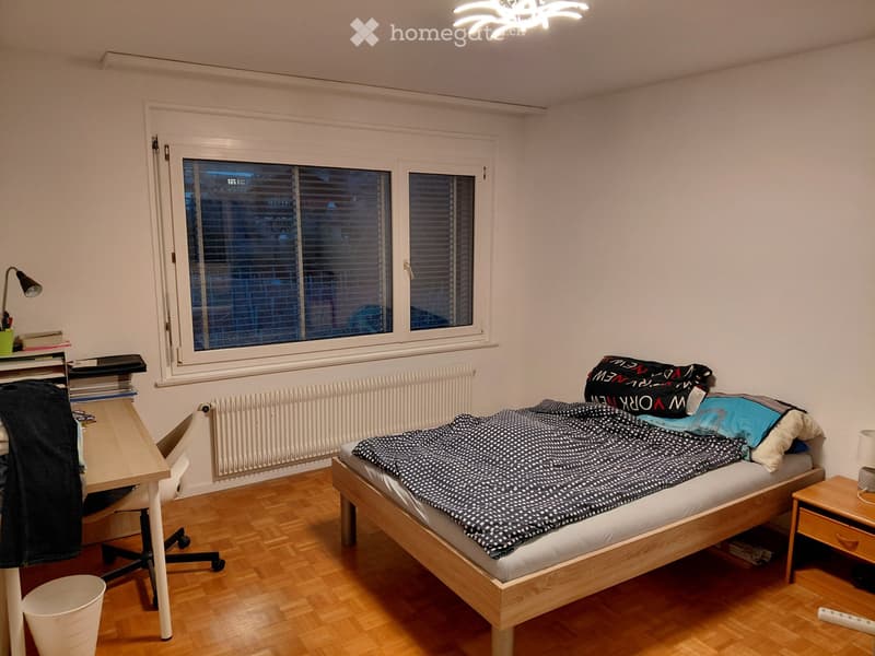 A tidy bedroom with a wooden bed, desk, chair, nightstand, and a window with blinds. The room features a wooden floor and white walls.
