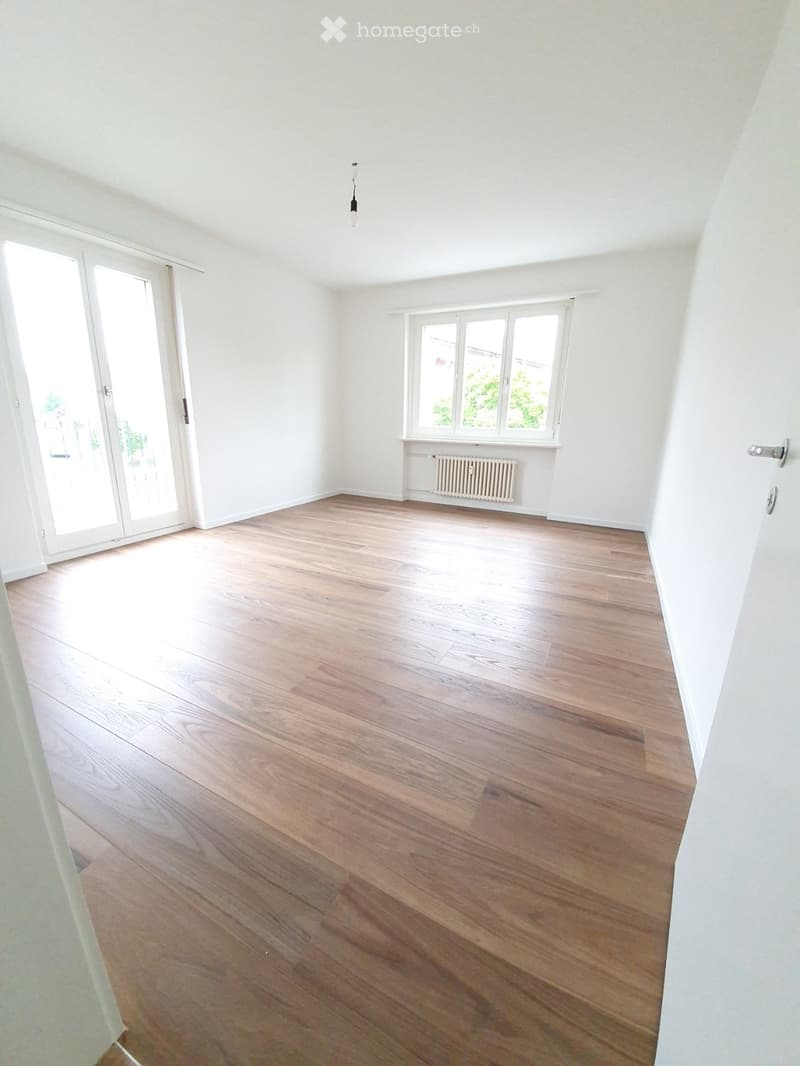 Empty room with light wooden flooring, white walls, large windows, and a sliding glass door leading outside.