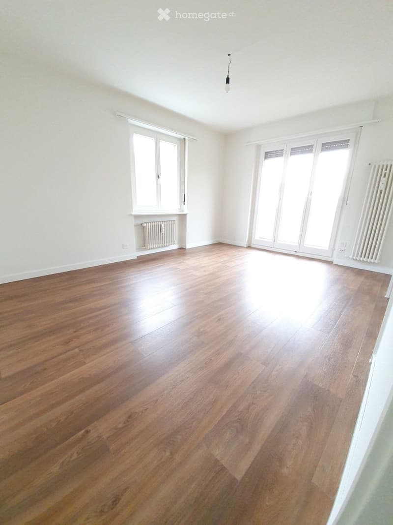 Empty room with wooden flooring, white walls, large windows, and a hanging light fixture.