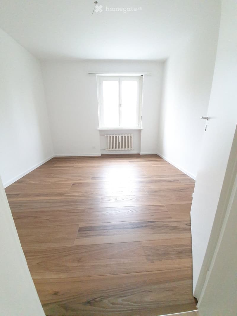 Empty room with wooden flooring, white walls, and a single window with a radiator below it.