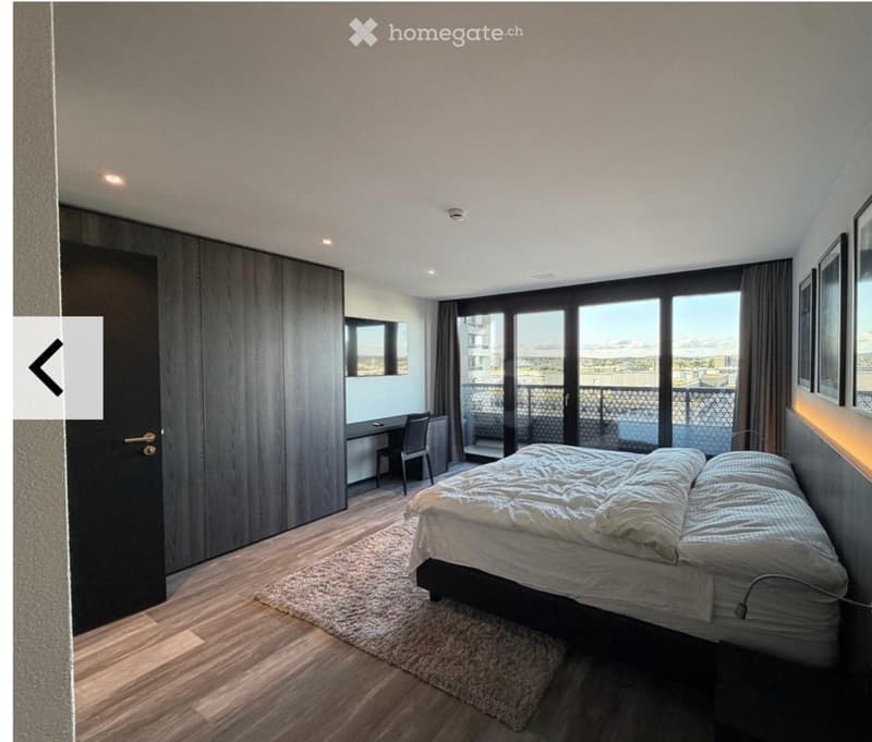 Modern bedroom with a large bed, wooden wardrobe, desk, chair, and large windows offering a view of the city.