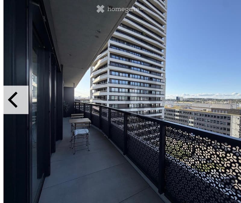 Balcony with a small table and chair, overlooking a modern high-rise building and cityscape.