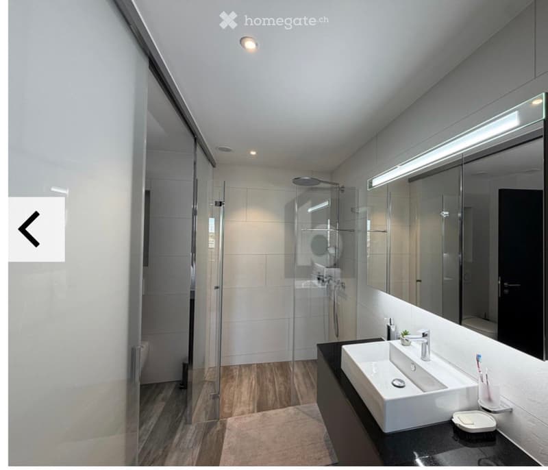 Modern bathroom with a white sink, large mirror with built-in lighting, glass-enclosed shower, and wooden flooring.