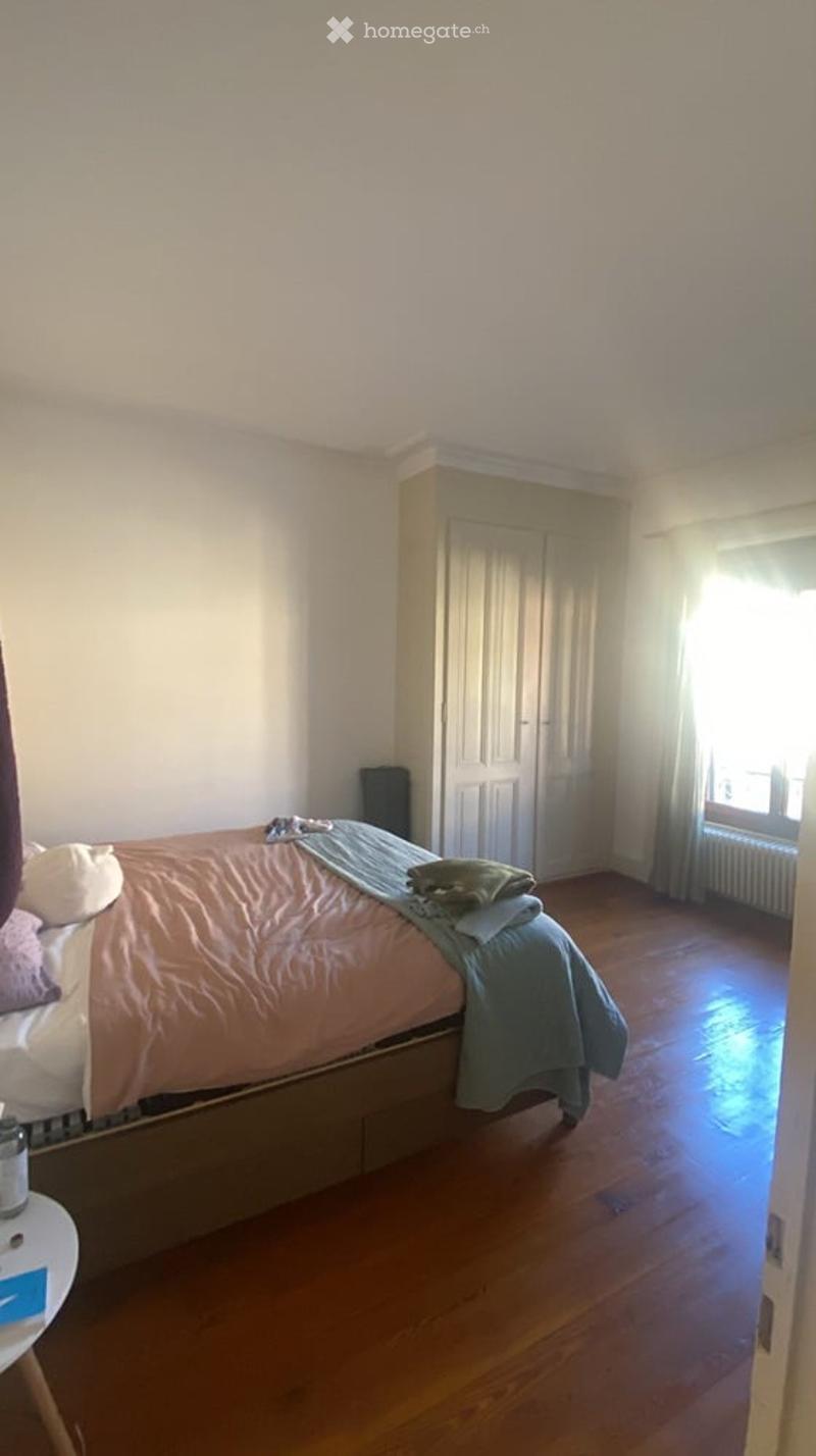 A cozy bedroom with wooden flooring, a bed with a peach-colored duvet and pillows, a nightstand, a wardrobe, and a window with curtains.