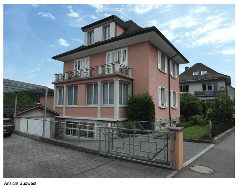 Two-story pink house with white trim, multiple windows, balconies, and a garage. The house is surrounded by greenery and a paved driveway.