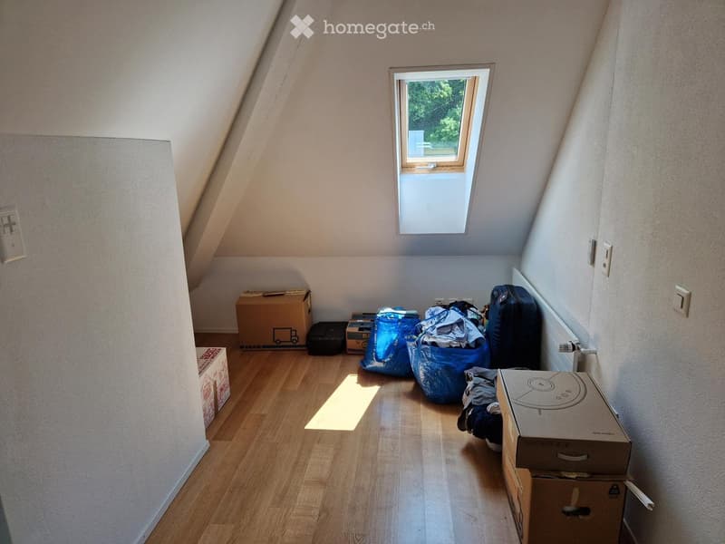 An attic room with wooden flooring, slanted ceilings, and a skylight window. The room is empty except for some boxes and bags, indicating it is being packed or unpacked.