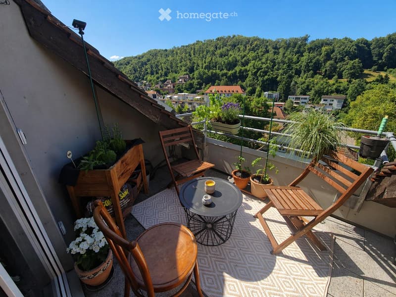 Balcony with wooden chairs, a small round table, and potted plants, offering a scenic view of a green, hilly landscape.