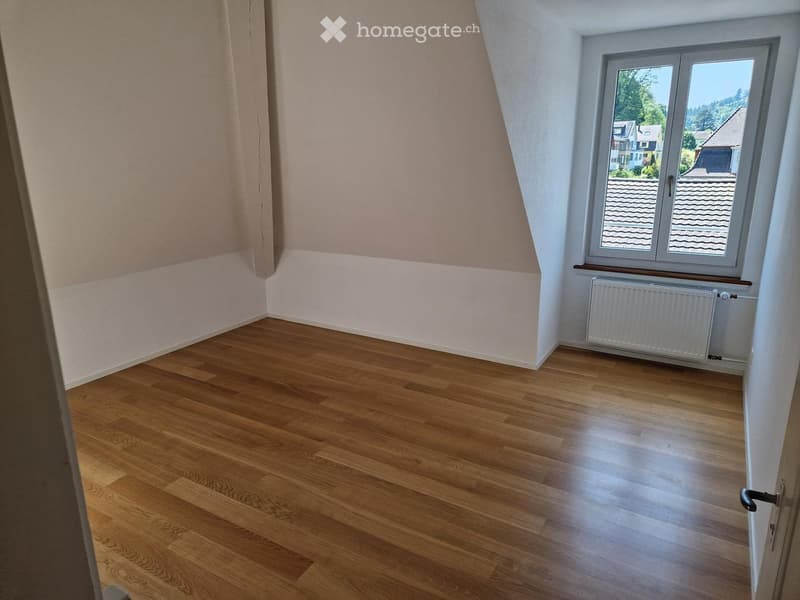 Empty attic room with wooden flooring, slanted ceilings, and a large window offering a view of neighboring rooftops.