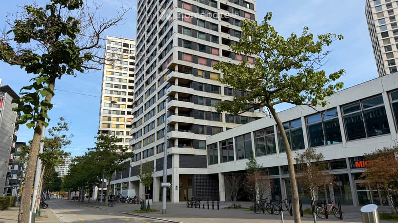 Modern multi-story residential building with balconies and large windows, surrounded by trees and parked bicycles.