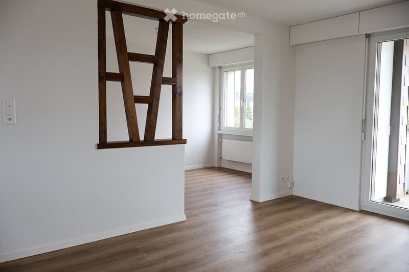 Empty room with light wooden flooring, white walls, and a large window with a view. A wooden shelf is mounted on the wall, and there is a sliding glass door leading to another space.