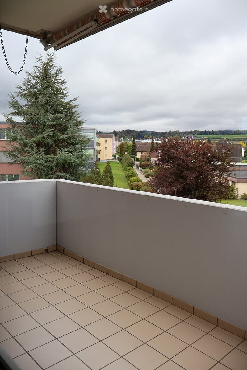 Balcony with tiled floor, low railing, and view of a residential area with trees and buildings.