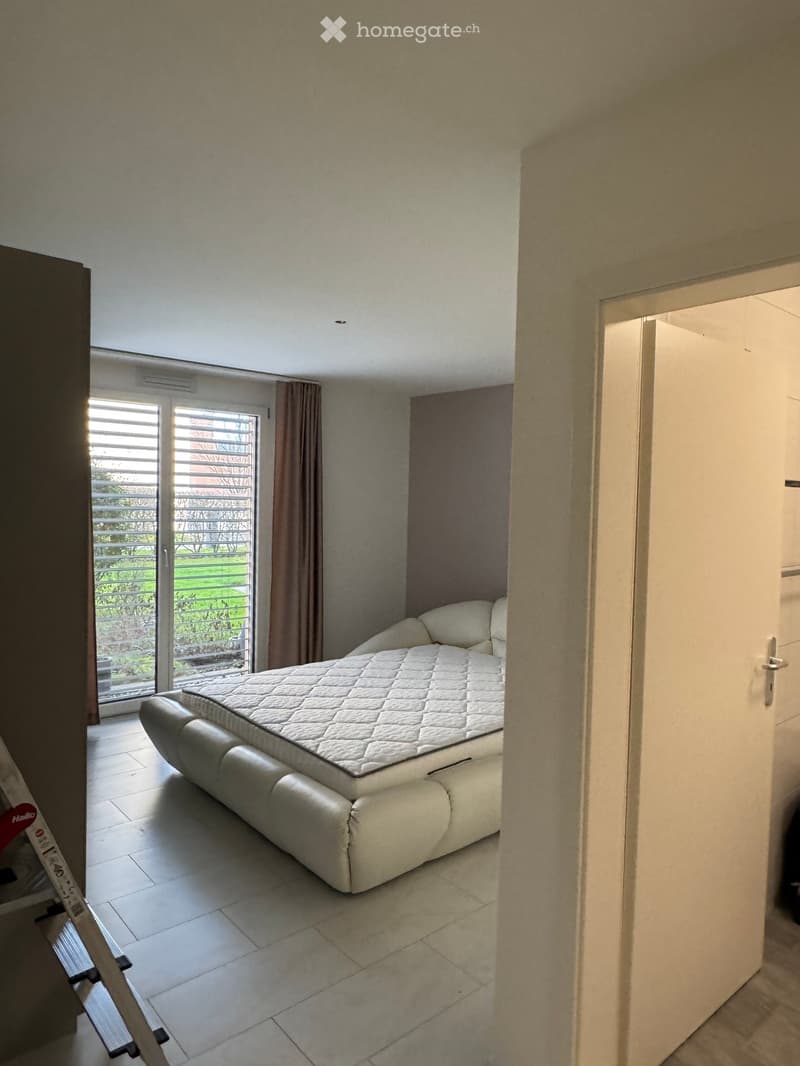 Modern bedroom with a white sofa bed, large window with blinds, and a view of a green outdoor area.