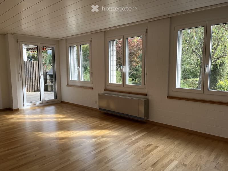 Empty room with wooden flooring, white brick walls, large windows, and a radiator. The room has a sliding glass door leading to an outdoor area.