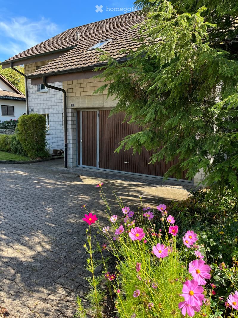 A two-story house with a tiled roof, white brick facade, and a wooden garage door. The driveway is paved, and there are pink flowers in the foreground.