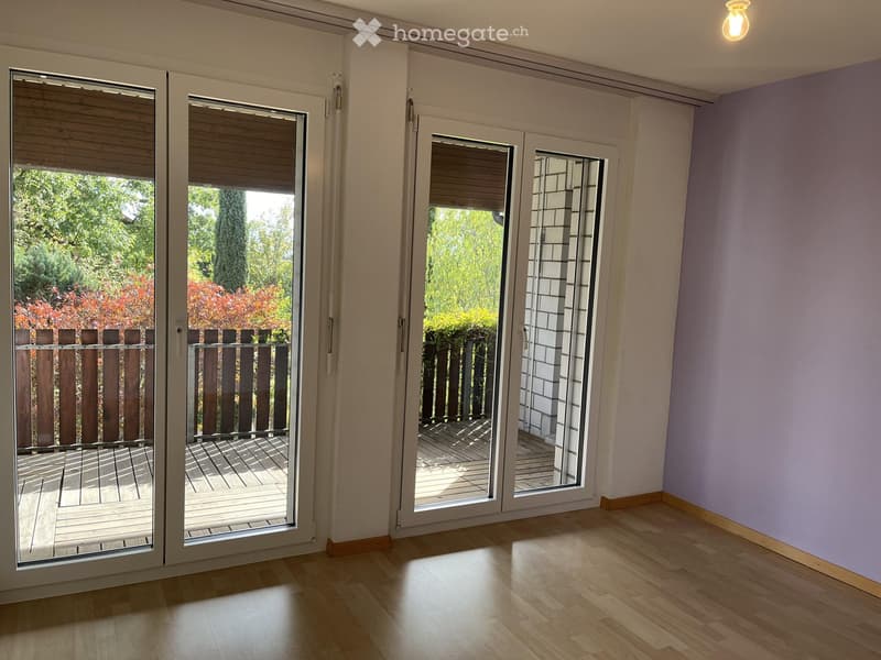 Empty room with wooden flooring, white walls, and large sliding glass doors leading to a wooden deck and garden area.