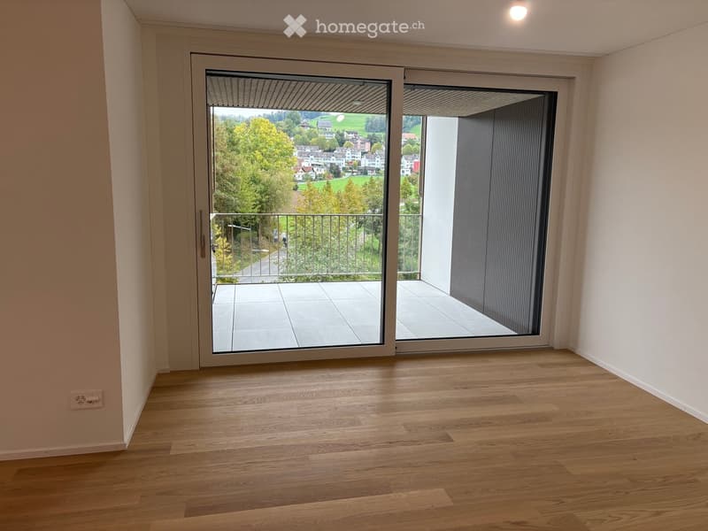 Empty room with wooden flooring, large sliding glass doors leading to a balcony with a scenic view of a town and greenery.