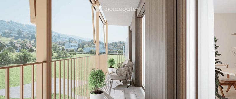 Balcony with a view of a green landscape and distant buildings, featuring a wicker chair, small table, and potted plants.