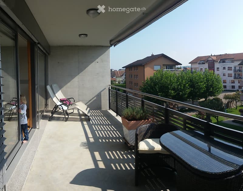 Balcony with a wicker chair, small table, potted plant, and lounge chairs, overlooking a residential area with houses and greenery.