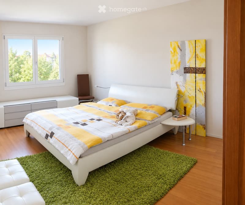 Modern bedroom with a white bed, yellow and grey bedding, green shag rug, white leather stool, brown chair, small round table with a book, and a colorful abstract art piece on the wall.