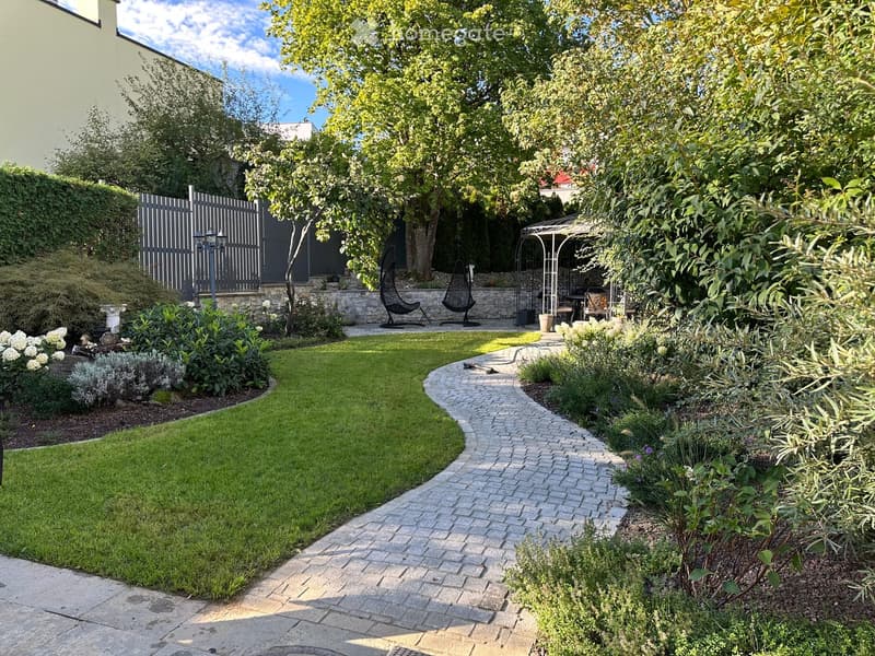 Well-maintained garden with a curved brick pathway, neatly trimmed lawn, various plants, trees, and outdoor seating area with hanging chairs and a small table.