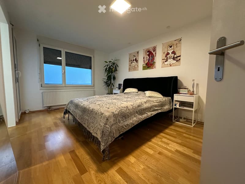 Spacious bedroom with wooden flooring, large window, bed with patterned bedspread, nightstand, plant, and artwork on walls.