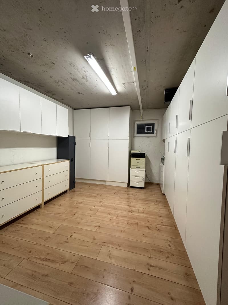 Empty room with light wooden flooring, white cabinetry, and a microwave mounted on the wall. The ceiling is concrete with exposed beams and a single fluorescent light fixture.