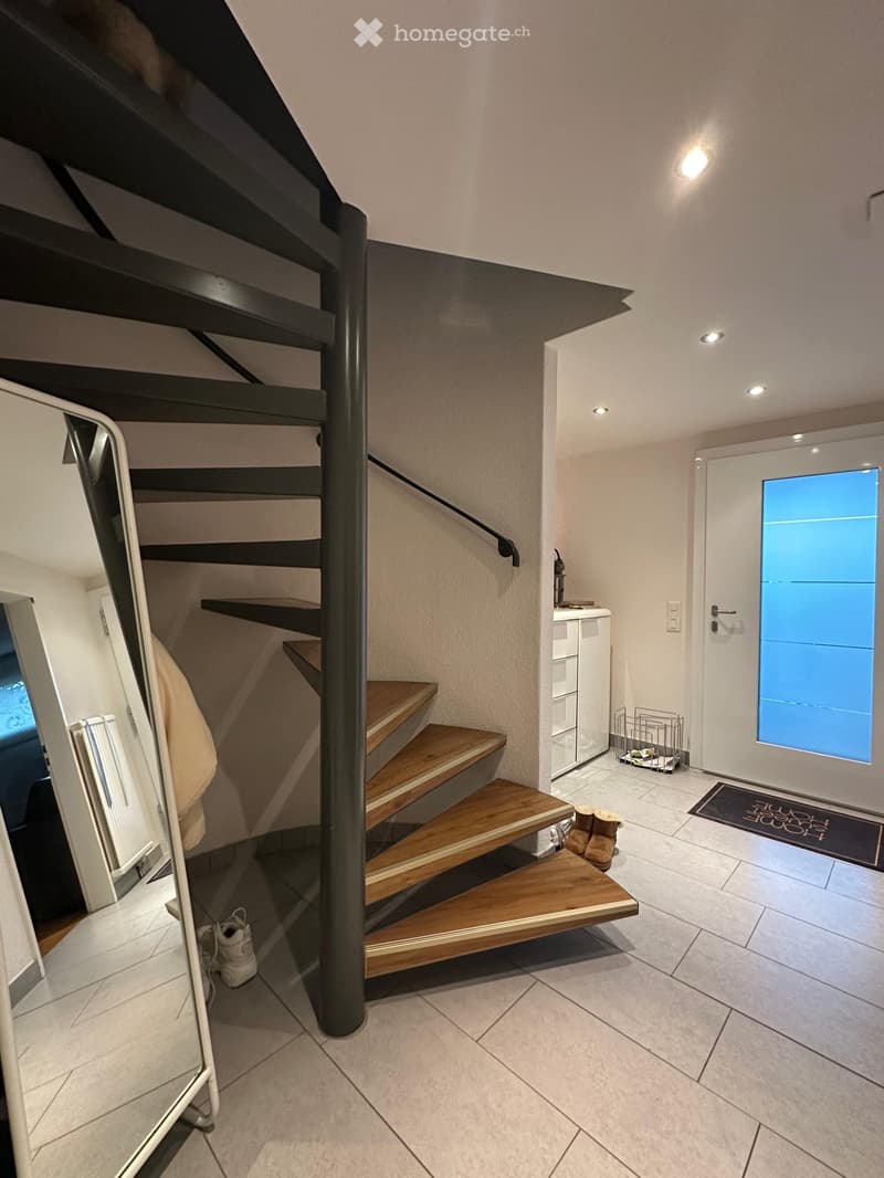 Modern hallway with a wooden staircase, white walls, and a small entryway area with a white door and a small cabinet.